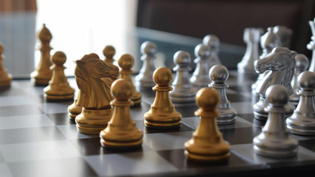 Dettaglio di una scacchiera con una partita già iniziata, le cui pedine sono di colore oro per un giocatore e di colore argento per l'altro.