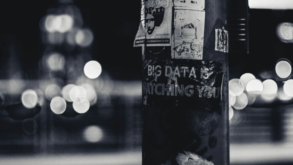 Un'immagine in bianco e nero con effetto Bokeh, con in primo piano una sezione di un palo della luce con attaccatto un adesivo che mostra le parole "big data is watching you"